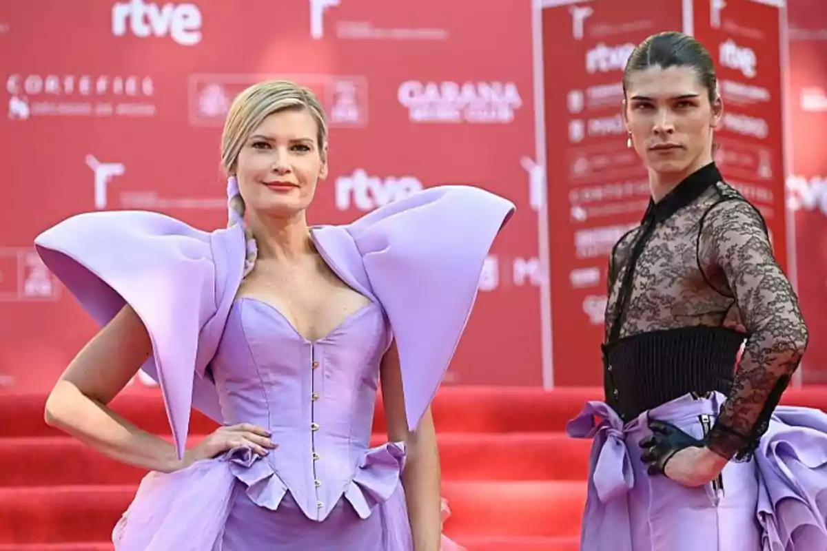 Dos personas posan en una alfombra roja con atuendos llamativos en tonos lila y detalles voluminosos, sobre un fondo rojo con logotipos de patrocinadores. Dos personas posan en una alfombra roja con atuendos llamativos en tonos lila y detalles voluminosos, sobre un fondo rojo con logotipos de patrocinadores.