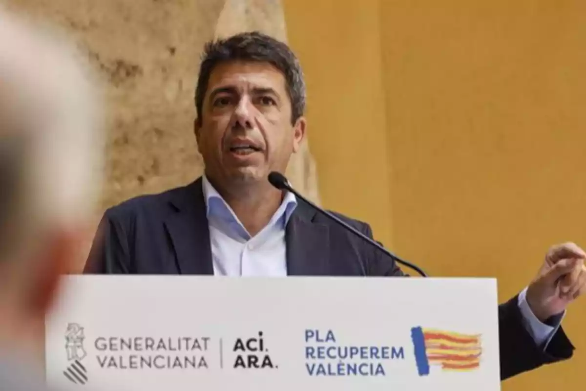 Hombre de cabello corto y oscuro hablando en un podio con un micrófono, detrás de un cartel que tiene los logotipos de la Generalitat Valenciana y el Pla Recuperem València, en un entorno interior con paredes de color beige. Hombre de cabello corto y oscuro hablando en un podio con un micrófono, detrás de un cartel que tiene los logotipos de la Generalitat Valenciana y el Pla Recuperem València, en un entorno interior con paredes de color beige.