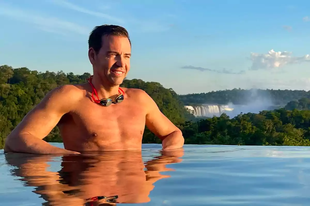 Hombre sonriente en una piscina con gafas de natación en el cuello y una cascada al fondo rodeada de vegetación