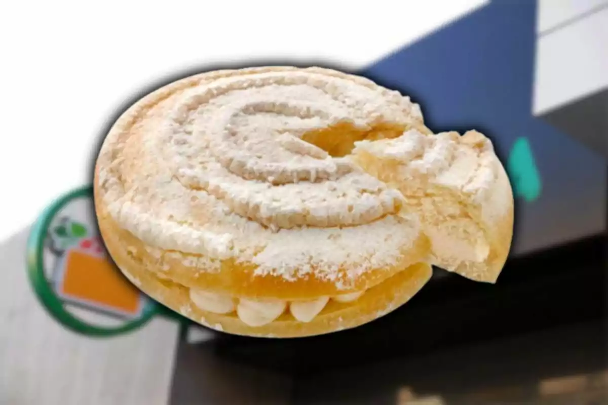 Un pan dulce tipo concha relleno de crema con una mordida, al fondo se observa la fachada de una tienda. Un pan dulce tipo concha relleno de crema con una mordida, al fondo se observa la fachada de una tienda.
