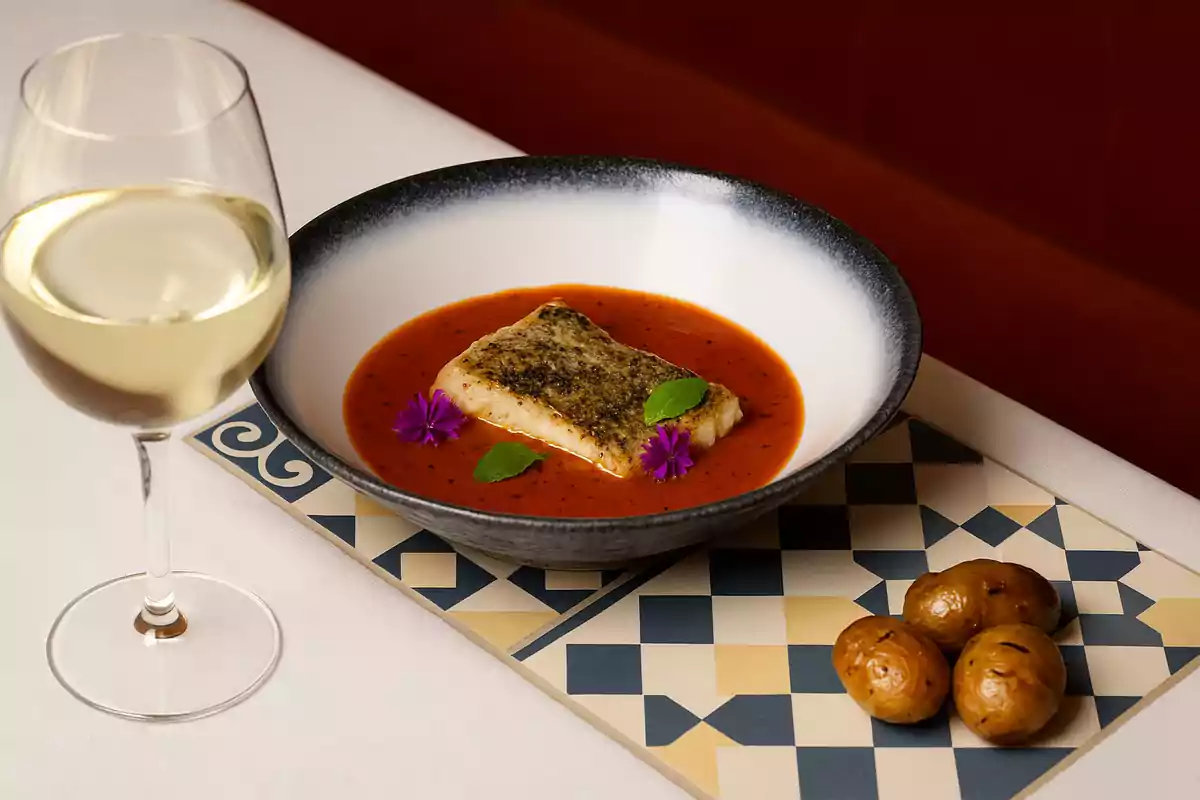 Plato de pescado con salsa roja decorado con flores y hojas verdes acompañado de papas asadas y una copa de vino blanco sobre una mesa elegante