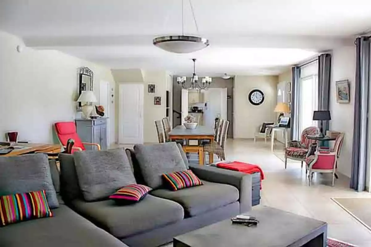 Sala y comedor modernos con sofá gris, cojines de colores, mesa de comedor de madera, sillas tejidas, lámparas colgantes y grandes ventanales con cortinas grises
