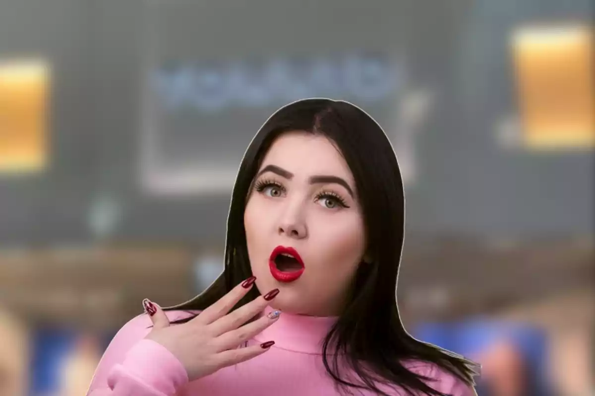 Mujer de cabello oscuro con expresión de sorpresa, usando suéter rosa y uñas largas rojas, fondo desenfocado