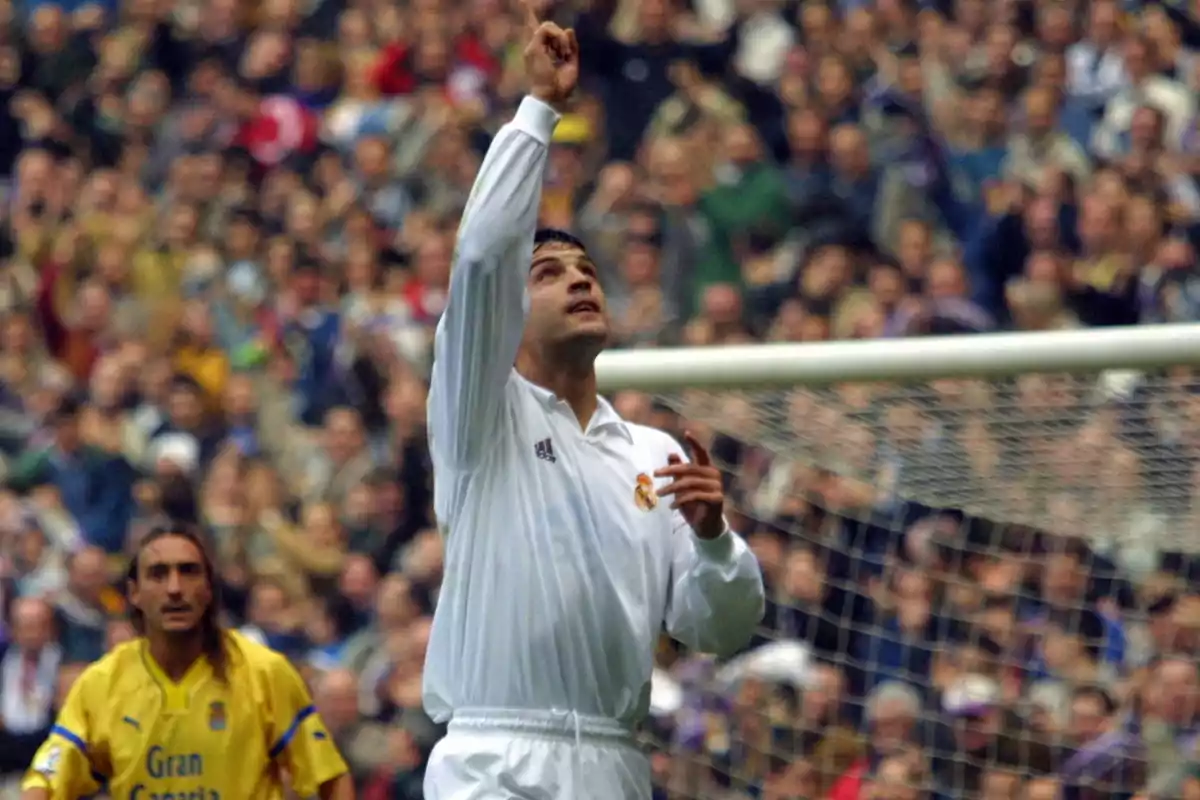 Futbolista del Real Madrid celebrando un gol señalando al cielo frente a la portería mientras un jugador rival observa Futbolista del Real Madrid celebrando un gol señalando al cielo frente a la portería mientras un jugador rival observa