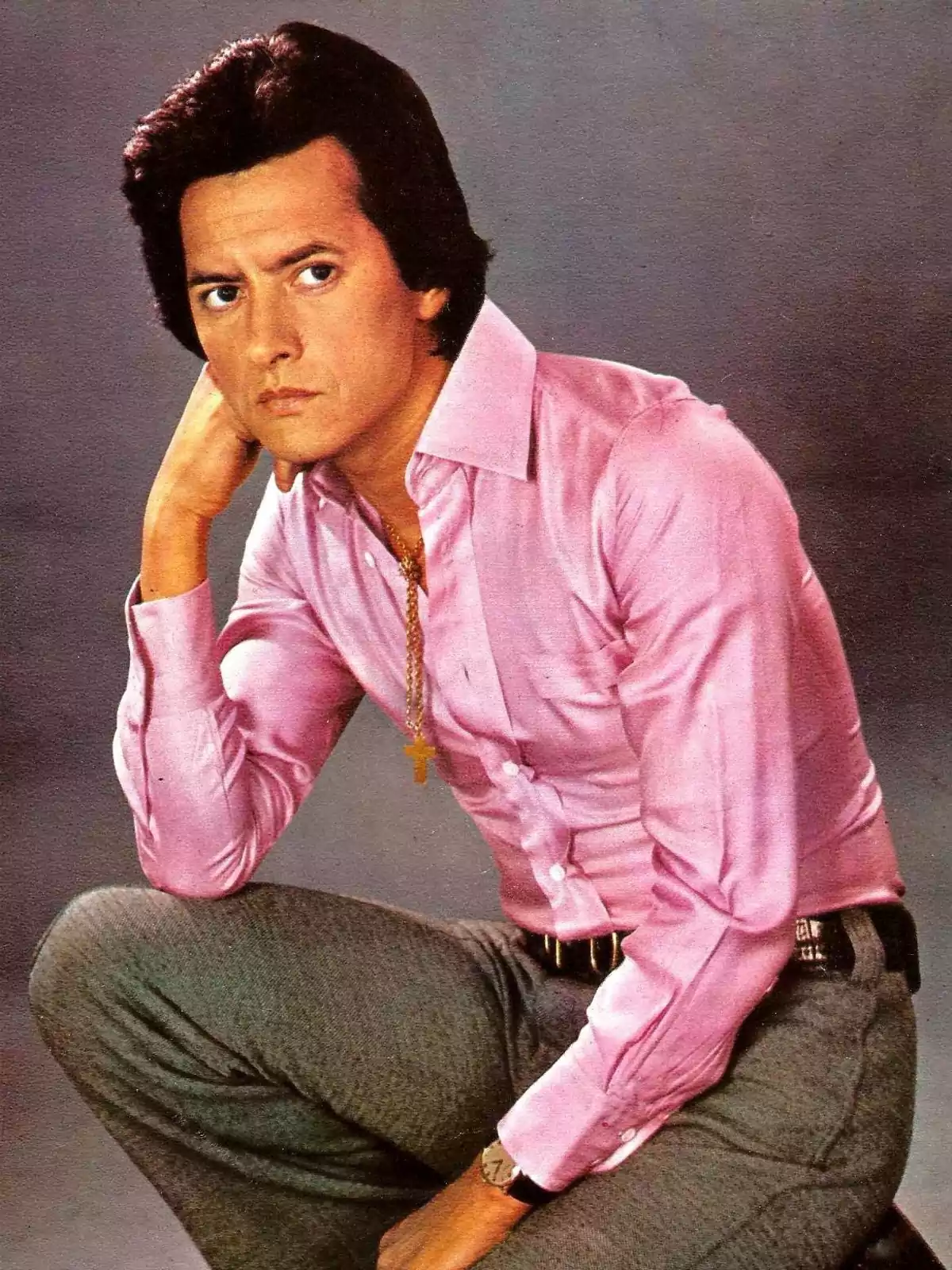 Hombre de cabello oscuro con camisa rosa y pantalón gris posando sentado con expresión seria