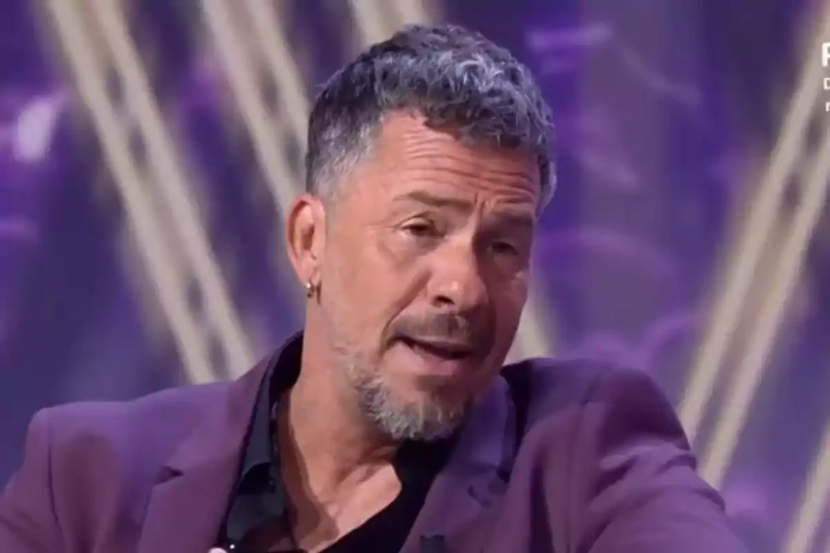 Hombre de cabello corto y canoso con barba y pendiente en la oreja, vestido con saco morado, hablando en un programa de televisión con fondo púrpura. Hombre de cabello corto y canoso con barba y pendiente en la oreja, vestido con saco morado, hablando en un programa de televisión con fondo púrpura.