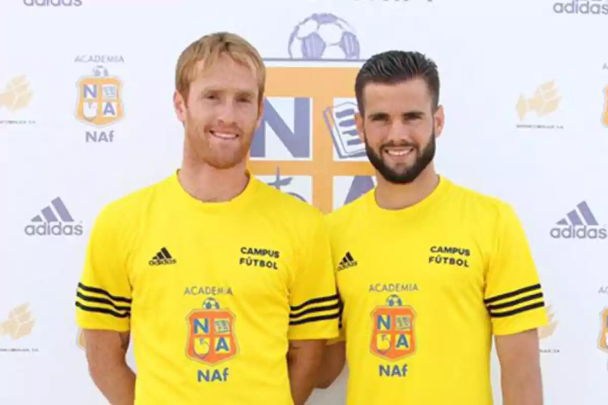 Dos hombres con camiseta amarilla de una academia de fútbol posan sonrientes frente a un fondo con logotipos deportivos. Dos hombres con camiseta amarilla de una academia de fútbol posan sonrientes frente a un fondo con logotipos deportivos.