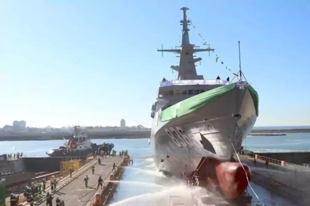 Un buque militar es botado al agua desde un astillero mientras varias personas observan y trabajan en el muelle