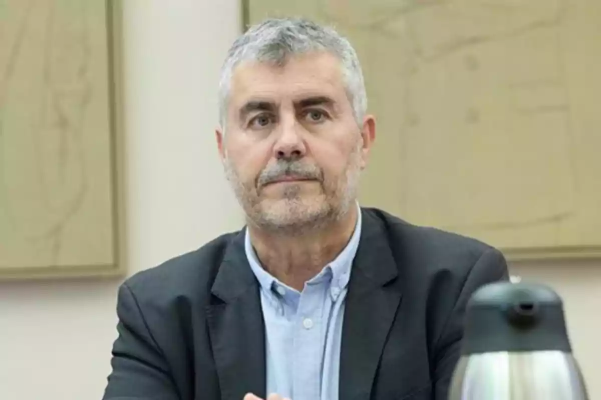Hombre de cabello canoso y barba corta con expresión seria, vestido con saco oscuro y camisa clara, sentado en una mesa con una cafetera plateada al frente y fondo de pared clara con cuadros