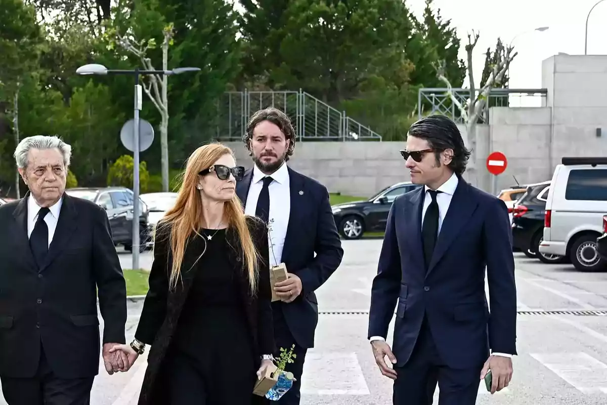 Cuatro personas vestidas de negro caminan juntas en un estacionamiento, aparentando estar en un evento formal o funeral. Cuatro personas vestidas de negro caminan juntas en un estacionamiento, aparentando estar en un evento formal o funeral.