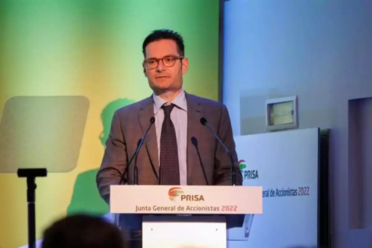 Hombre con gafas y traje hablando en un podio durante la Junta General de Accionistas 2022 de PRISA con fondo verde y amarillo. Hombre con gafas y traje hablando en un podio durante la Junta General de Accionistas 2022 de PRISA con fondo verde y amarillo.