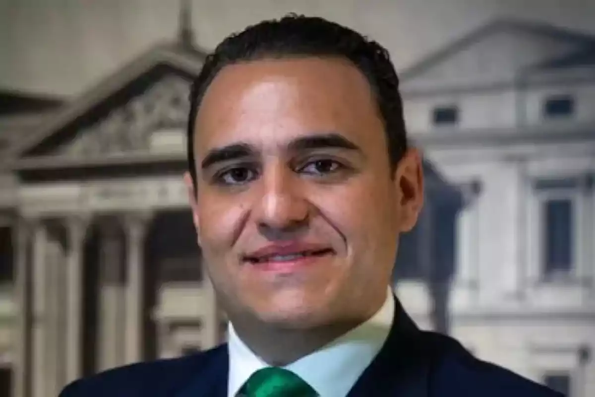 Hombre de cabello corto y oscuro con traje y corbata verde sonriendo frente a un fondo de edificio antiguo desenfocado Hombre de cabello corto y oscuro con traje y corbata verde sonriendo frente a un fondo de edificio antiguo desenfocado