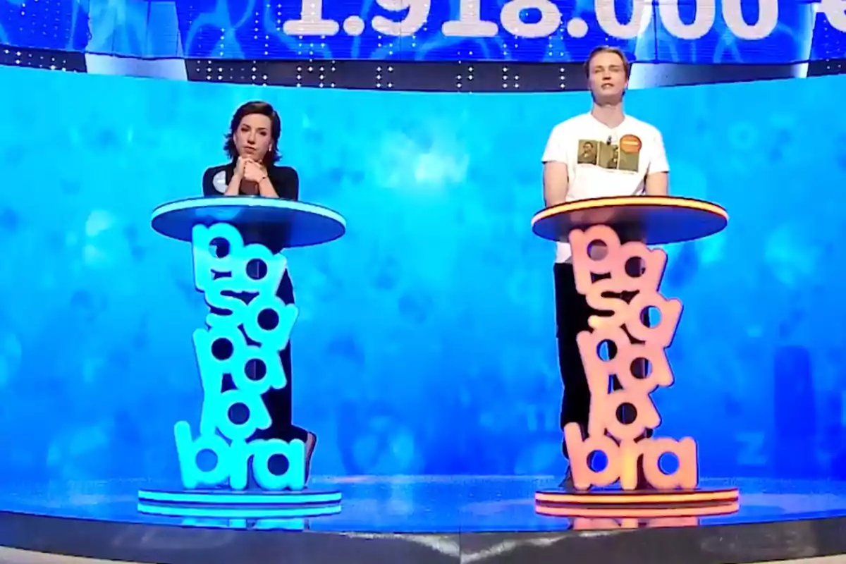 Rosa y Manu están de pie detrás de atriles coloridos en un set de televisión con fondo azul, participando en un programa de juegos. Captura de 'Pasapalabra' Rosa y Manu están de pie detrás de atriles coloridos en un set de televisión con fondo azul, participando en un programa de juegos. Captura de 'Pasapalabra'