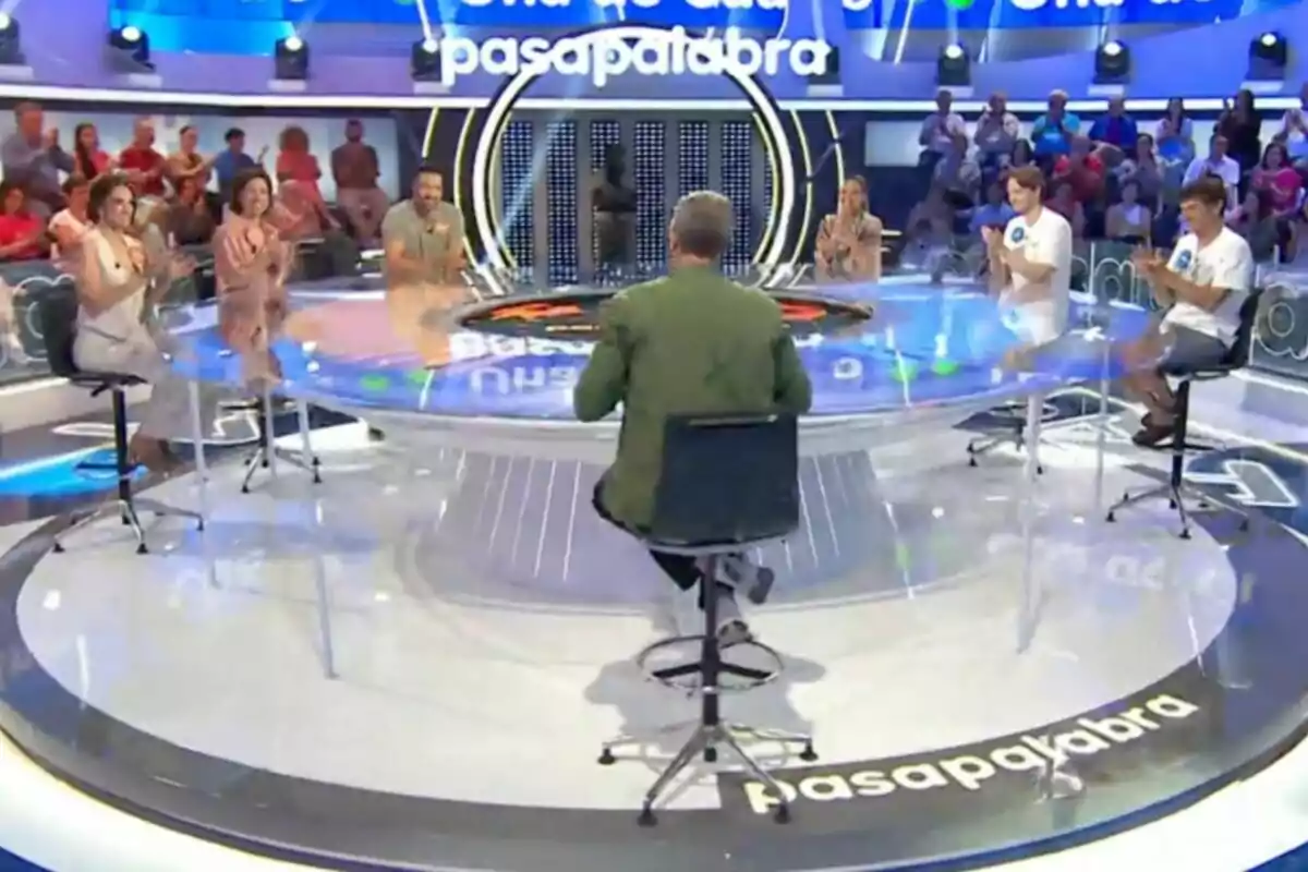 Personas sentadas alrededor de una mesa circular en un set de televisión con público al fondo y el logo de Pasapalabra en el suelo Personas sentadas alrededor de una mesa circular en un set de televisión con público al fondo y el logo de Pasapalabra en el suelo