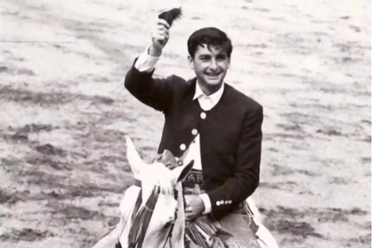 Joven sonriente vestido de traje corto montado a caballo en una plaza de toros levantando un trofeo en la mano Joven sonriente vestido de traje corto montado a caballo en una plaza de toros levantando un trofeo en la mano