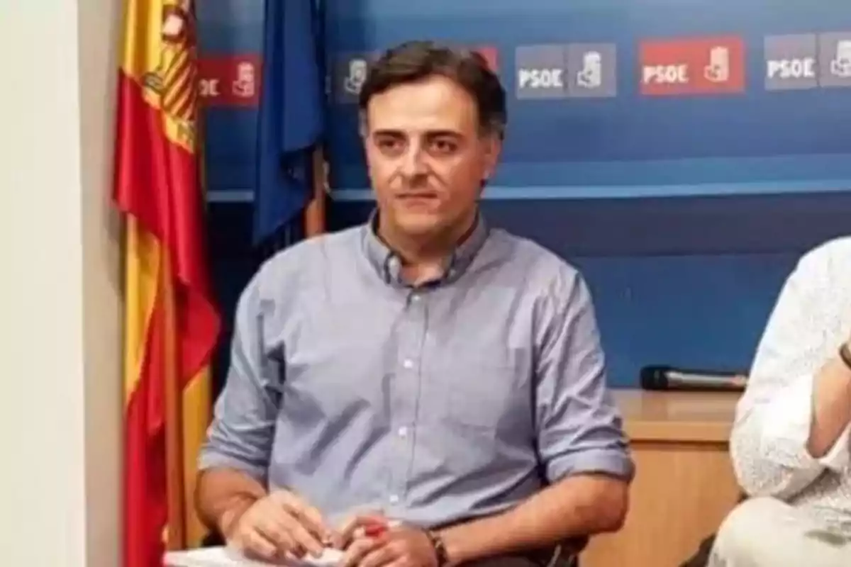 Hombre sentado con camisa azul claro frente a una pared azul con logotipos de PSOE y una bandera de España a su lado Hombre sentado con camisa azul claro frente a una pared azul con logotipos de PSOE y una bandera de España a su lado