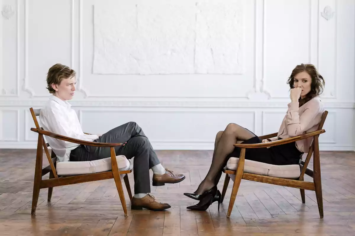 Dos personas sentadas una frente a la otra en sillas de madera en una habitación luminosa con suelo de madera y paredes blancas Dos personas sentadas una frente a la otra en sillas de madera en una habitación luminosa con suelo de madera y paredes blancas