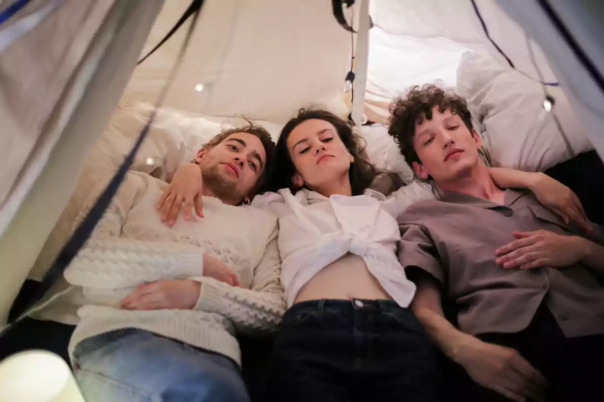 Tres personas jóvenes descansan juntas en el interior de una tienda de campaña improvisada con luces decorativas Tres personas jóvenes descansan juntas en el interior de una tienda de campaña improvisada con luces decorativas