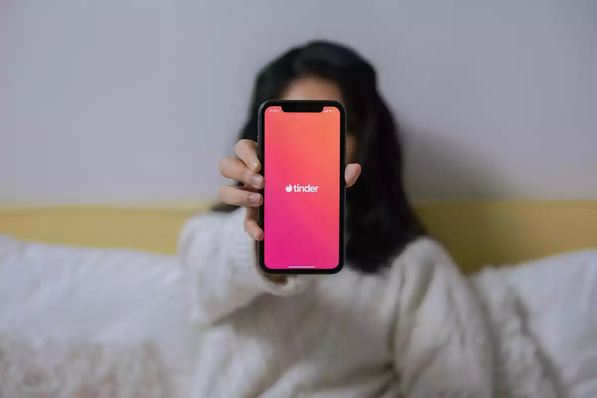 Persona sentada en una cama mostrando un teléfono móvil con la pantalla de inicio de la aplicación Tinder Persona sentada en una cama mostrando un teléfono móvil con la pantalla de inicio de la aplicación Tinder