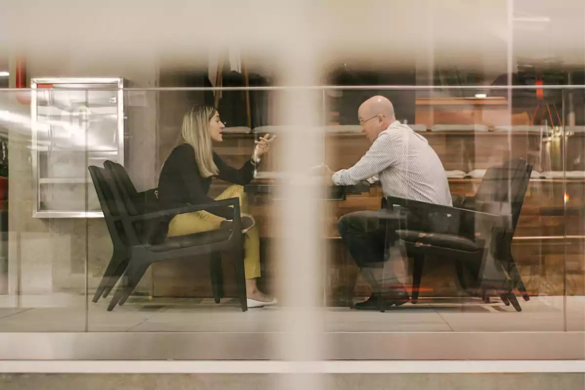 Dos personas sentadas frente a frente conversando en una mesa dentro de un espacio moderno con paredes de vidrio