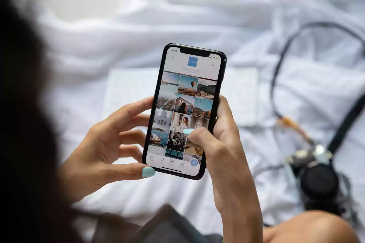 Persona usando un teléfono móvil para ver una galería de fotos de viajes mientras está sentada en una cama con una cámara y un teclado cerca Persona usando un teléfono móvil para ver una galería de fotos de viajes mientras está sentada en una cama con una cámara y un teclado cerca