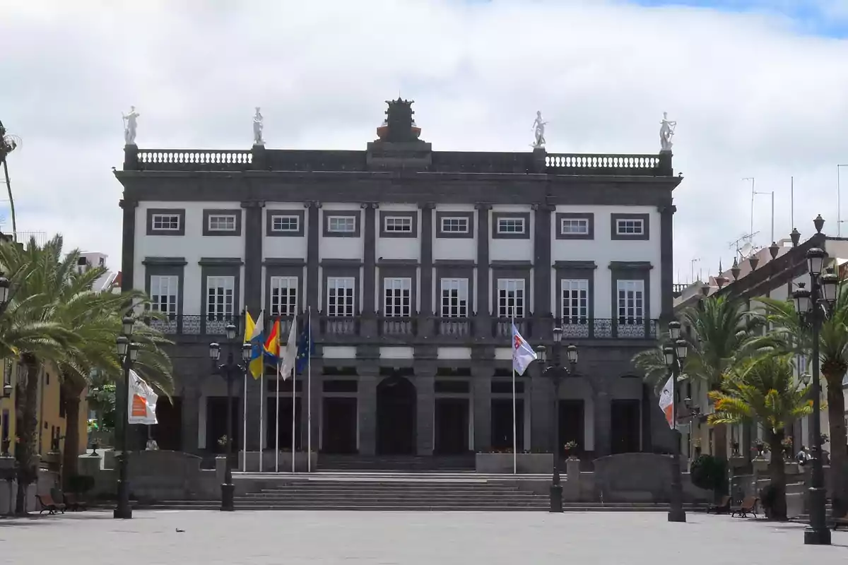 Fachada de un edificio histórico con columnas y estatuas en la parte superior rodeado de palmeras y banderas en una plaza amplia Fachada de un edificio histórico con columnas y estatuas en la parte superior rodeado de palmeras y banderas en una plaza amplia