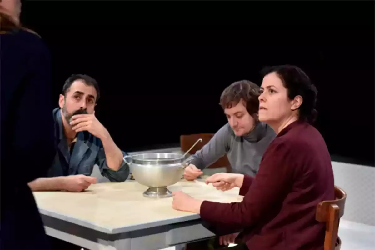 Tres personas sentadas alrededor de una mesa miran atentamente hacia alguien de pie mientras tienen platos y una sopera metálica frente a ellos Tres personas sentadas alrededor de una mesa miran atentamente hacia alguien de pie mientras tienen platos y una sopera metálica frente a ellos