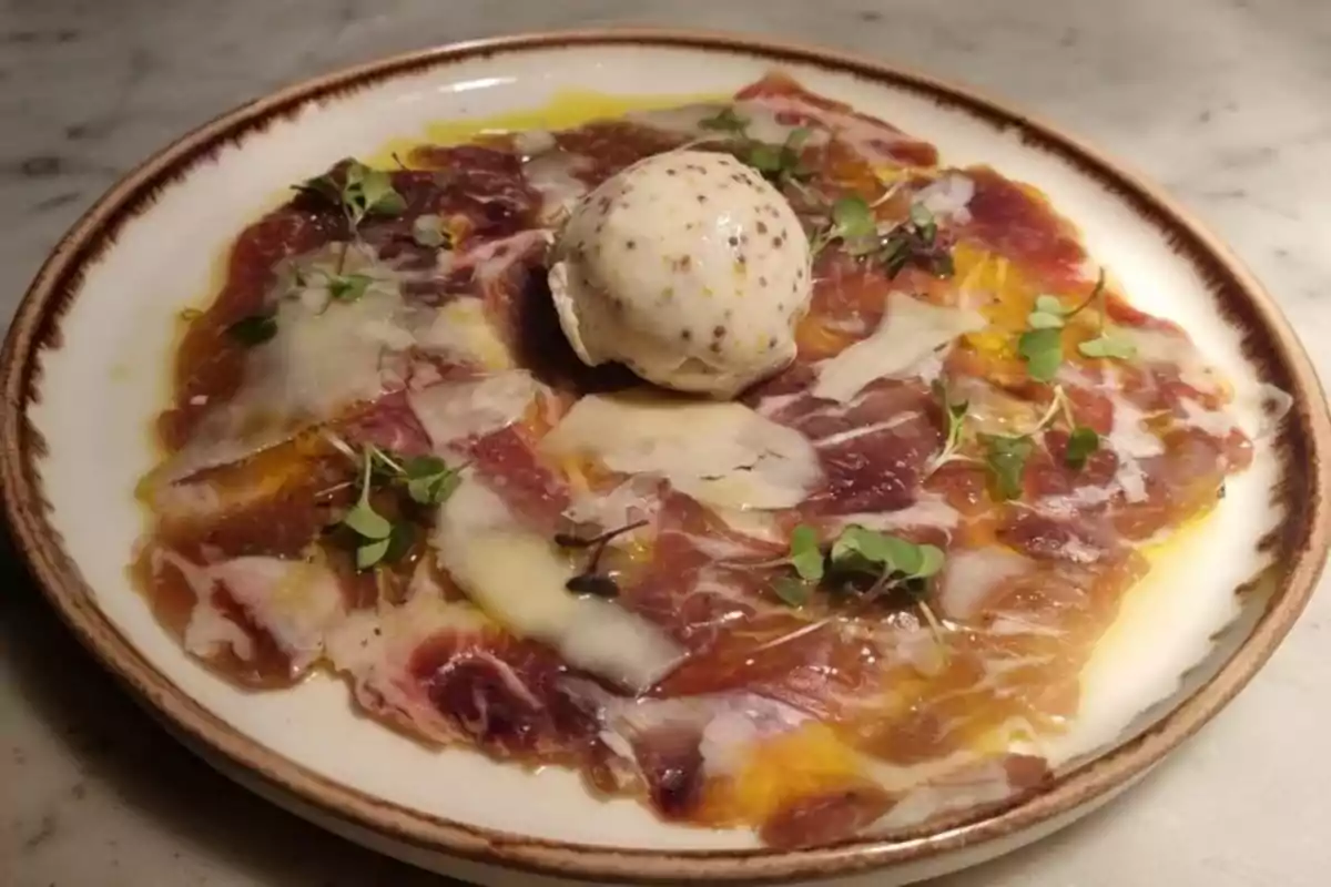 Plato de carpaccio de carne con lascas de queso, brotes verdes y una bola de helado o mousse en el centro, servido sobre un plato de cerámica. Plato de carpaccio de carne con lascas de queso, brotes verdes y una bola de helado o mousse en el centro, servido sobre un plato de cerámica.