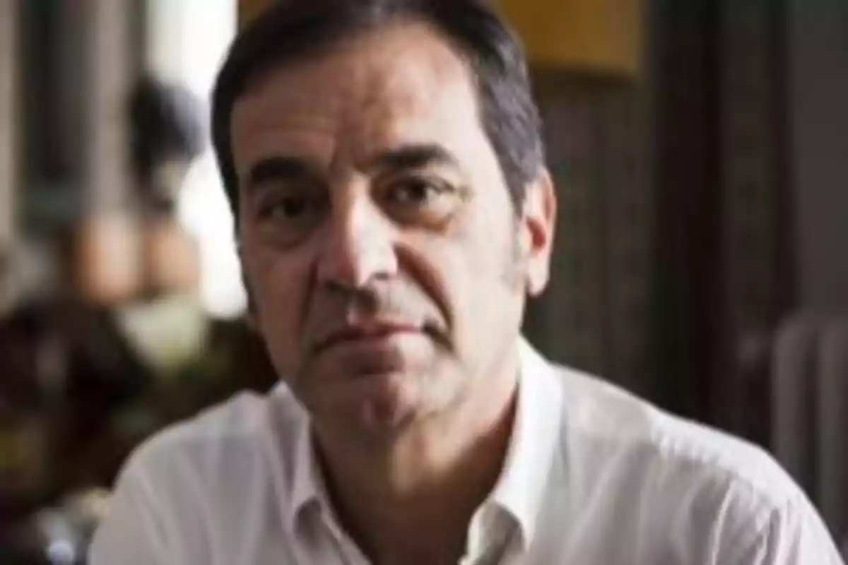 Hombre de mediana edad con camisa blanca mirando a la cámara en un ambiente interior desenfocado