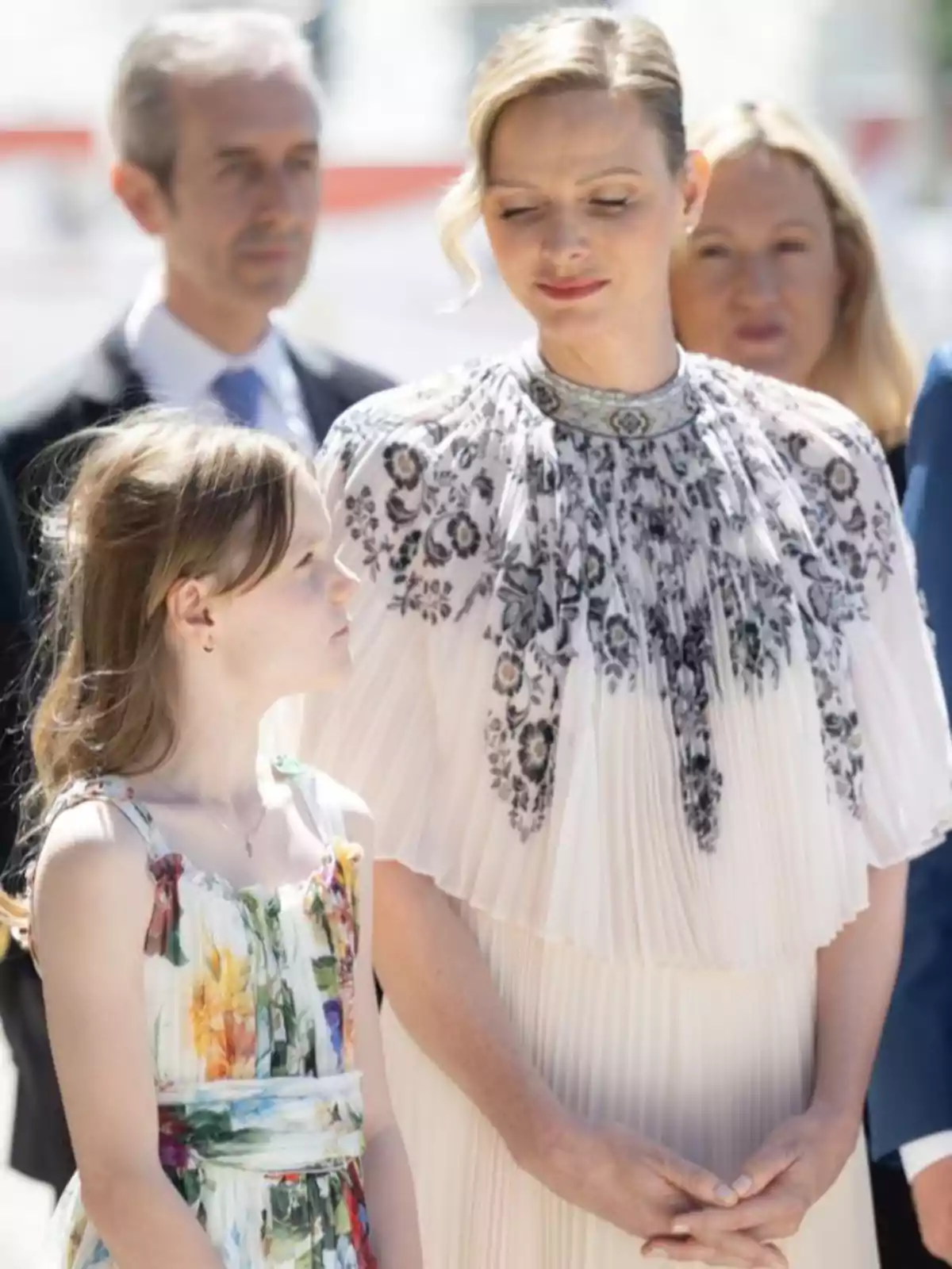Charlene con un vestido blanco con detalles florales en negro mira con ternura a Gabriella que lleva un vestido colorido, ambas están al aire libre y hay personas desenfocadas al fondo Charlene con un vestido blanco con detalles florales en negro mira con ternura a Gabriella que lleva un vestido colorido, ambas están al aire libre y hay personas desenfocadas al fondo