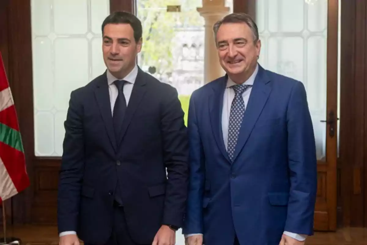 Dos hombres con traje posan sonrientes en una oficina elegante con una bandera en el fondo.