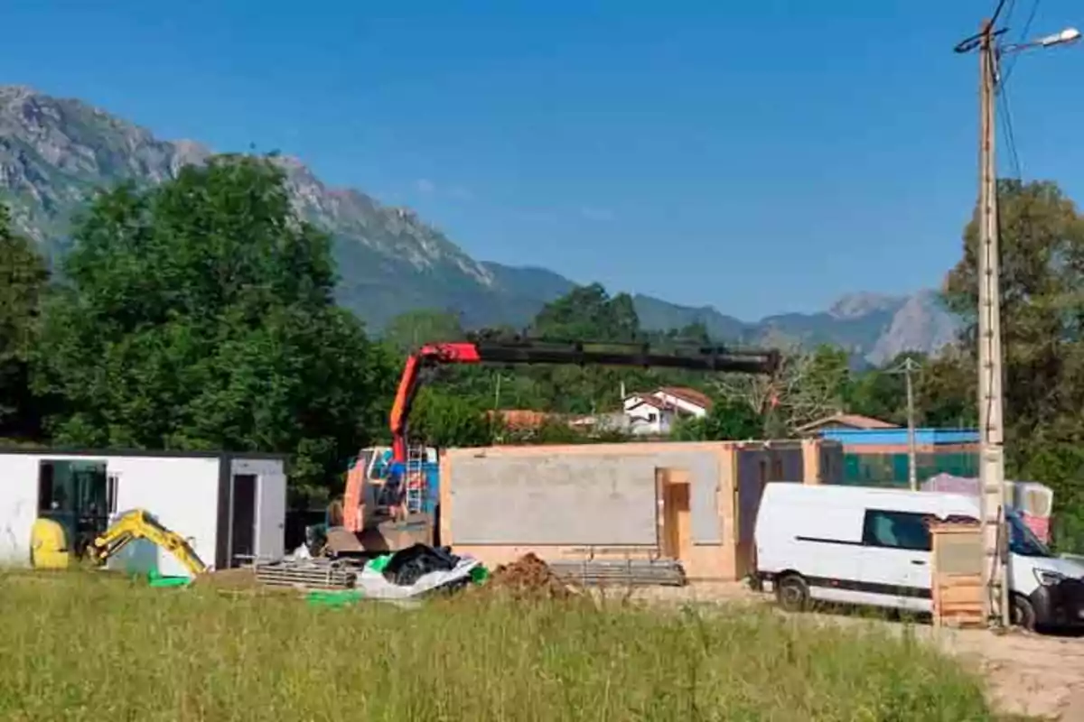 Obra en construcción en una zona rural con montañas al fondo, una grúa, una excavadora pequeña y una furgoneta blanca estacionada Obra en construcción en una zona rural con montañas al fondo, una grúa, una excavadora pequeña y una furgoneta blanca estacionada