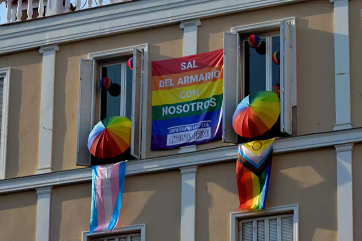 Fachada de edificio con dos ventanas abiertas decoradas con paraguas de colores y banderas LGBT y trans, además de una pancarta que dice Sal del armario con nosotros Fachada de edificio con dos ventanas abiertas decoradas con paraguas de colores y banderas LGBT y trans, además de una pancarta que dice Sal del armario con nosotros