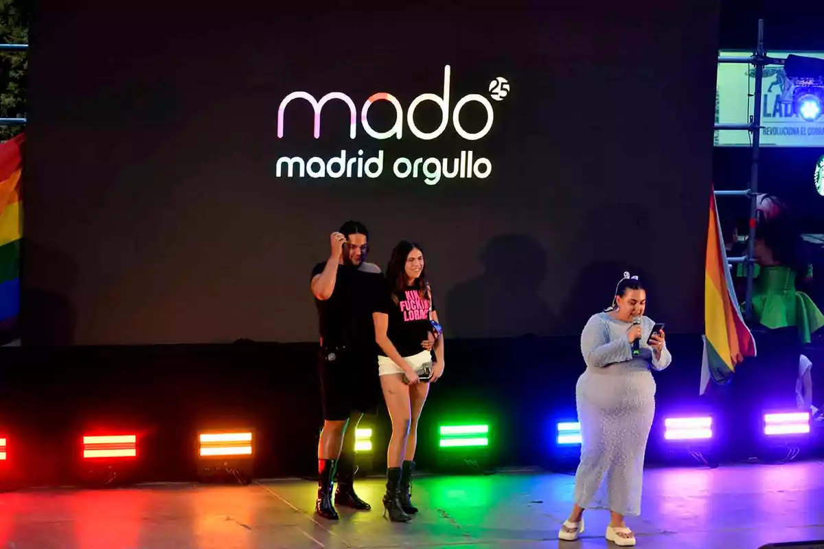 Tres personas en un escenario durante un evento de Madrid Orgullo con luces de colores y banderas arcoíris Tres personas en un escenario durante un evento de Madrid Orgullo con luces de colores y banderas arcoíris
