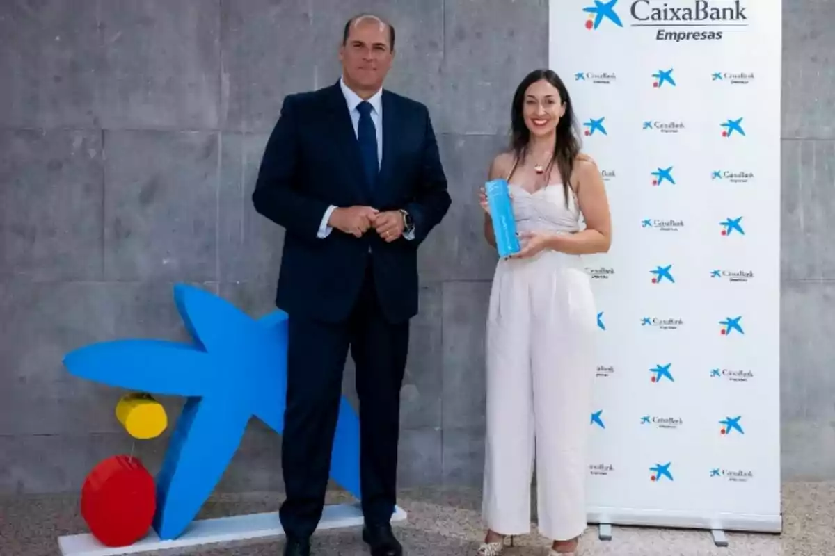 Un hombre con traje oscuro y una mujer con un mono blanco posan sonrientes en un evento de CaixaBank Empresas, ella sostiene un trofeo azul y ambos están frente a un fondo gris y un cartel corporativo Un hombre con traje oscuro y una mujer con un mono blanco posan sonrientes en un evento de CaixaBank Empresas, ella sostiene un trofeo azul y ambos están frente a un fondo gris y un cartel corporativo