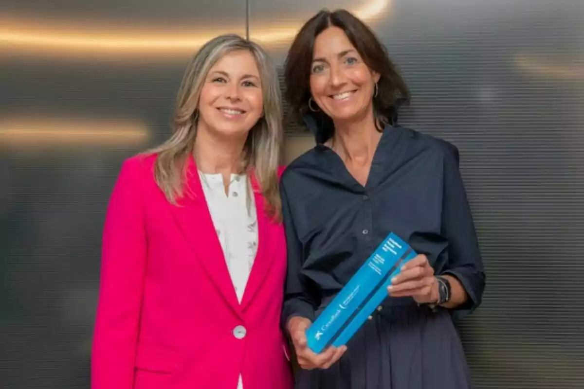 Dos mujeres sonríen a la cámara, una lleva un saco fucsia y la otra una blusa azul oscuro mientras sostiene un objeto rectangular azul Dos mujeres sonríen a la cámara, una lleva un saco fucsia y la otra una blusa azul oscuro mientras sostiene un objeto rectangular azul