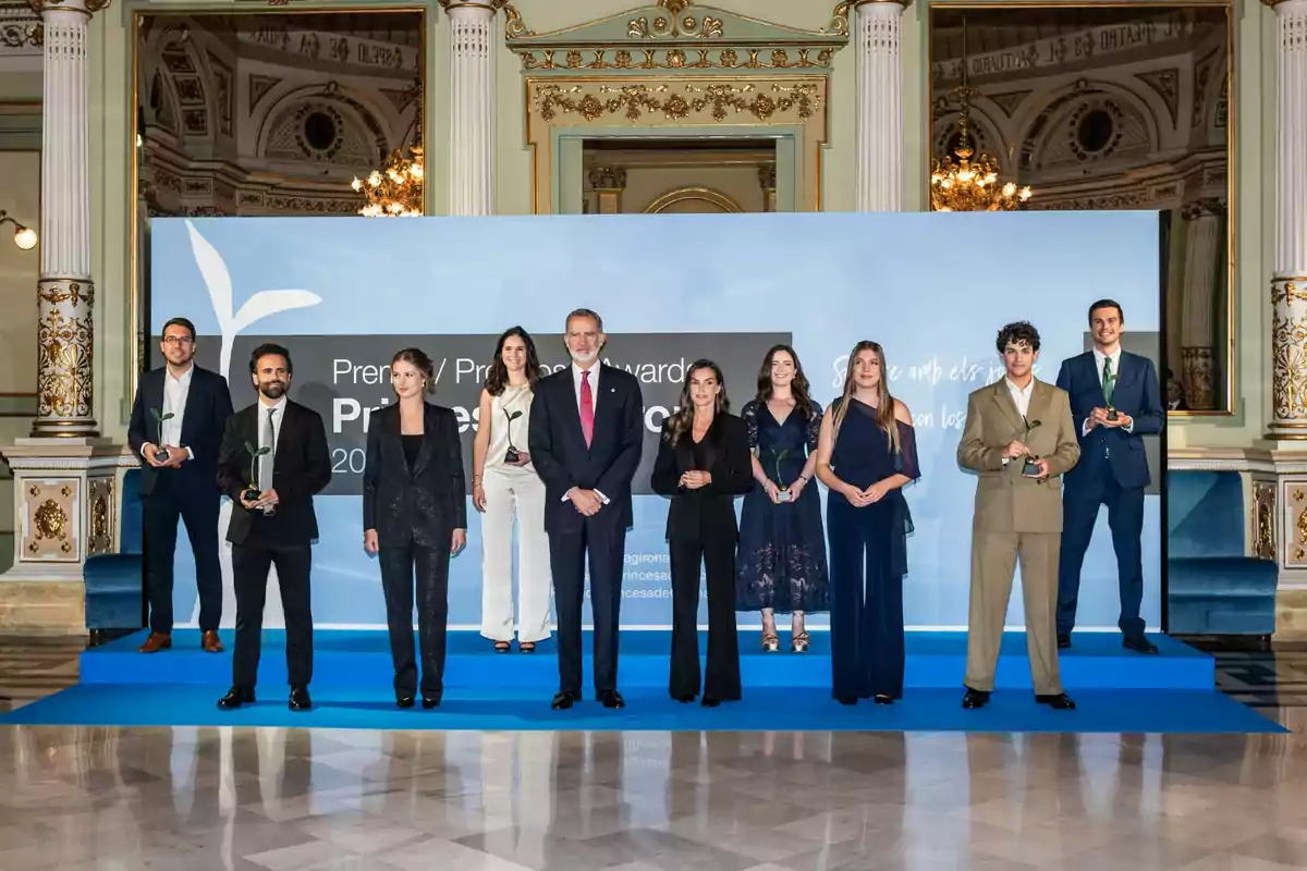 Un grupo de personas elegantemente vestidas posa sobre una tarima azul en un salón decorado de manera clásica y lujosa, sosteniendo trofeos en una ceremonia de premiación
