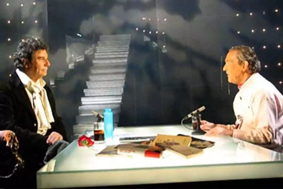 Dos hombres conversan sentados frente a frente en un set de televisión con una mesa de cristal, libros y bebidas, al fondo se ve una escalera iluminada y luces decorativas Dos hombres conversan sentados frente a frente en un set de televisión con una mesa de cristal, libros y bebidas, al fondo se ve una escalera iluminada y luces decorativas