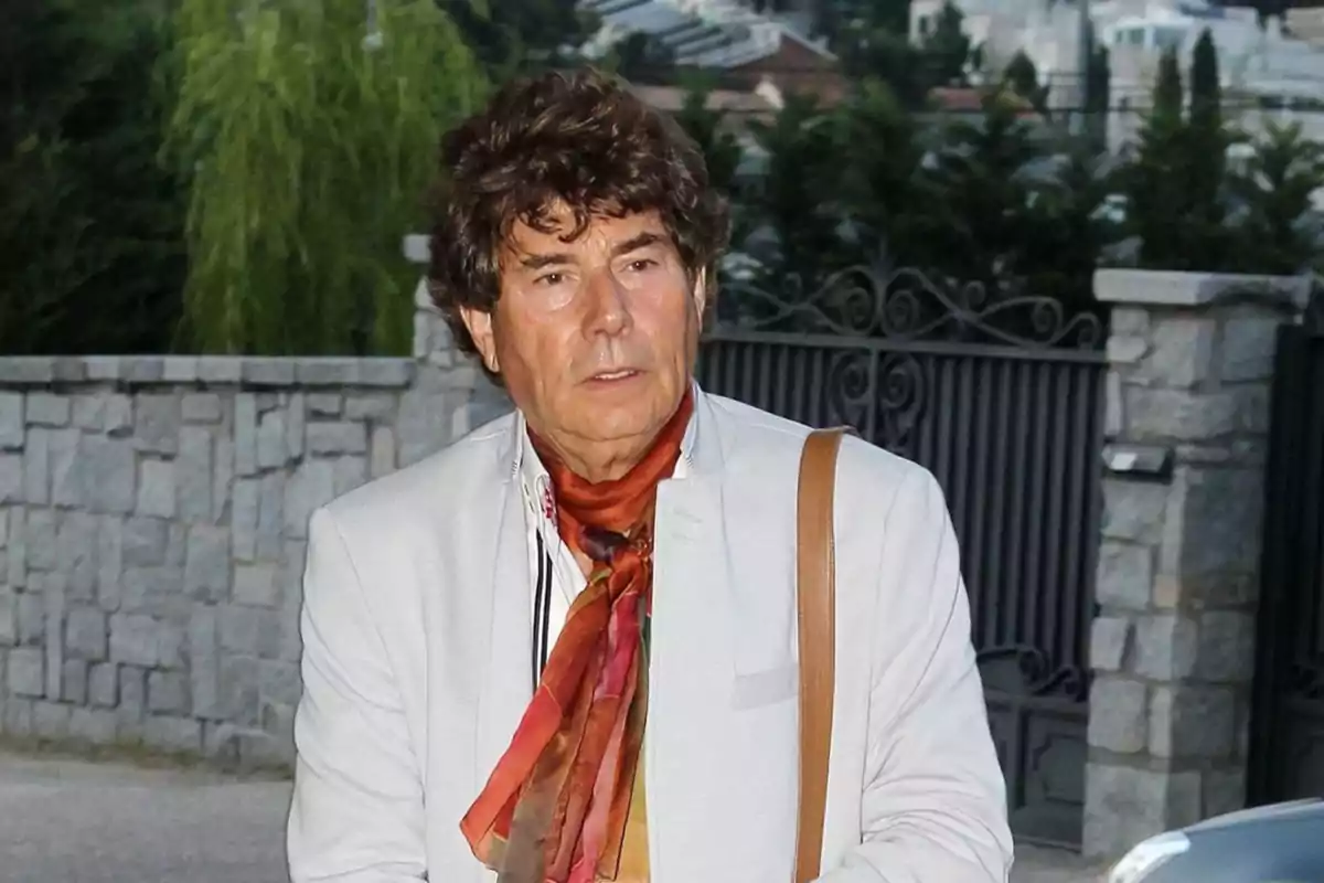 Hombre de cabello rizado y oscuro vistiendo saco claro y bufanda colorida, de pie frente a una reja y un muro de piedra en un entorno exterior