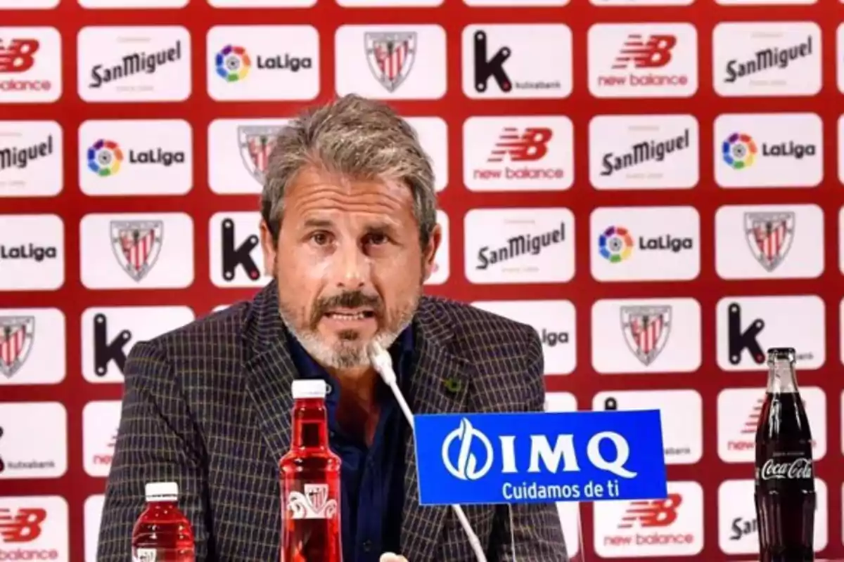 Hombre de cabello canoso y barba hablando en una rueda de prensa con fondo de logotipos de patrocinadores y botellas sobre la mesa