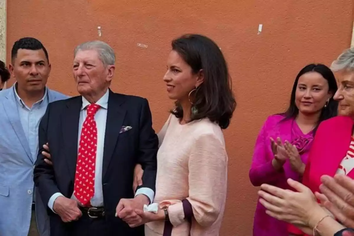 Un grupo de personas elegantemente vestidas posa frente a una pared naranja, algunas aplauden y otras miran hacia el frente Un grupo de personas elegantemente vestidas posa frente a una pared naranja, algunas aplauden y otras miran hacia el frente