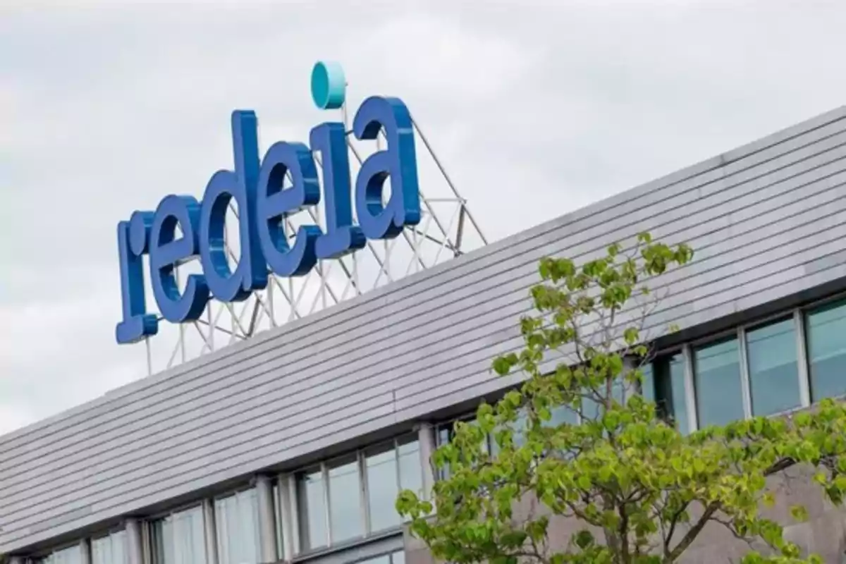 Edificio moderno con el logotipo de redeia en la parte superior y un árbol en primer plano Edificio moderno con el logotipo de redeia en la parte superior y un árbol en primer plano