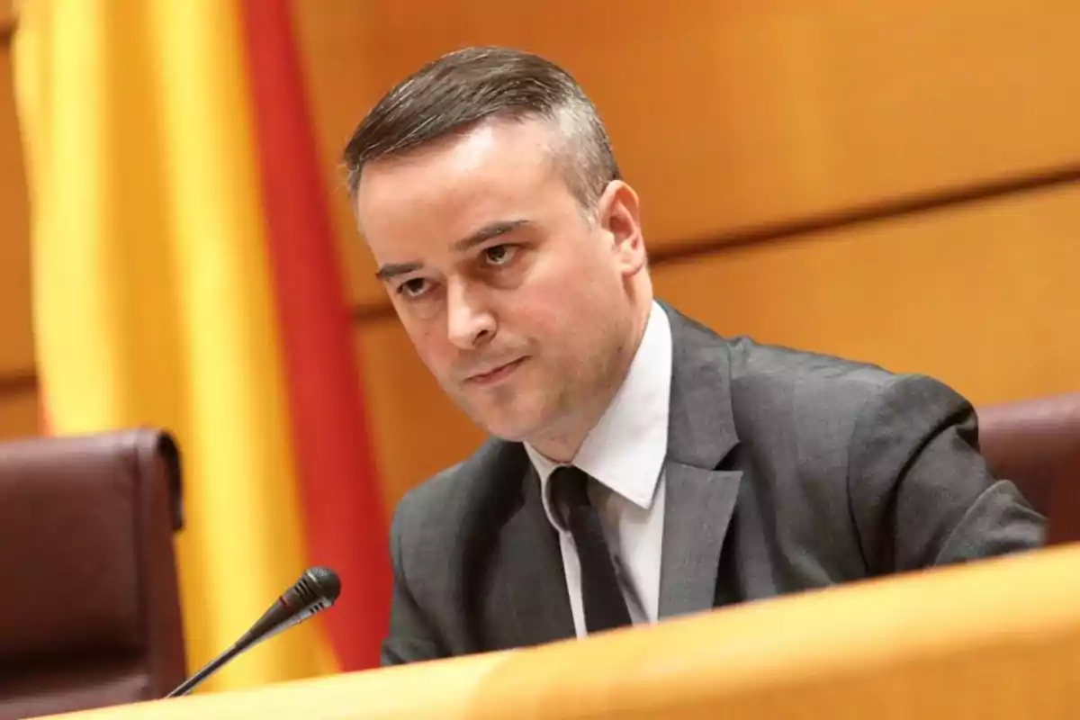 Hombre de traje gris sentado en una sala de conferencias con una bandera de fondo y un micrófono frente a él Hombre de traje gris sentado en una sala de conferencias con una bandera de fondo y un micrófono frente a él
