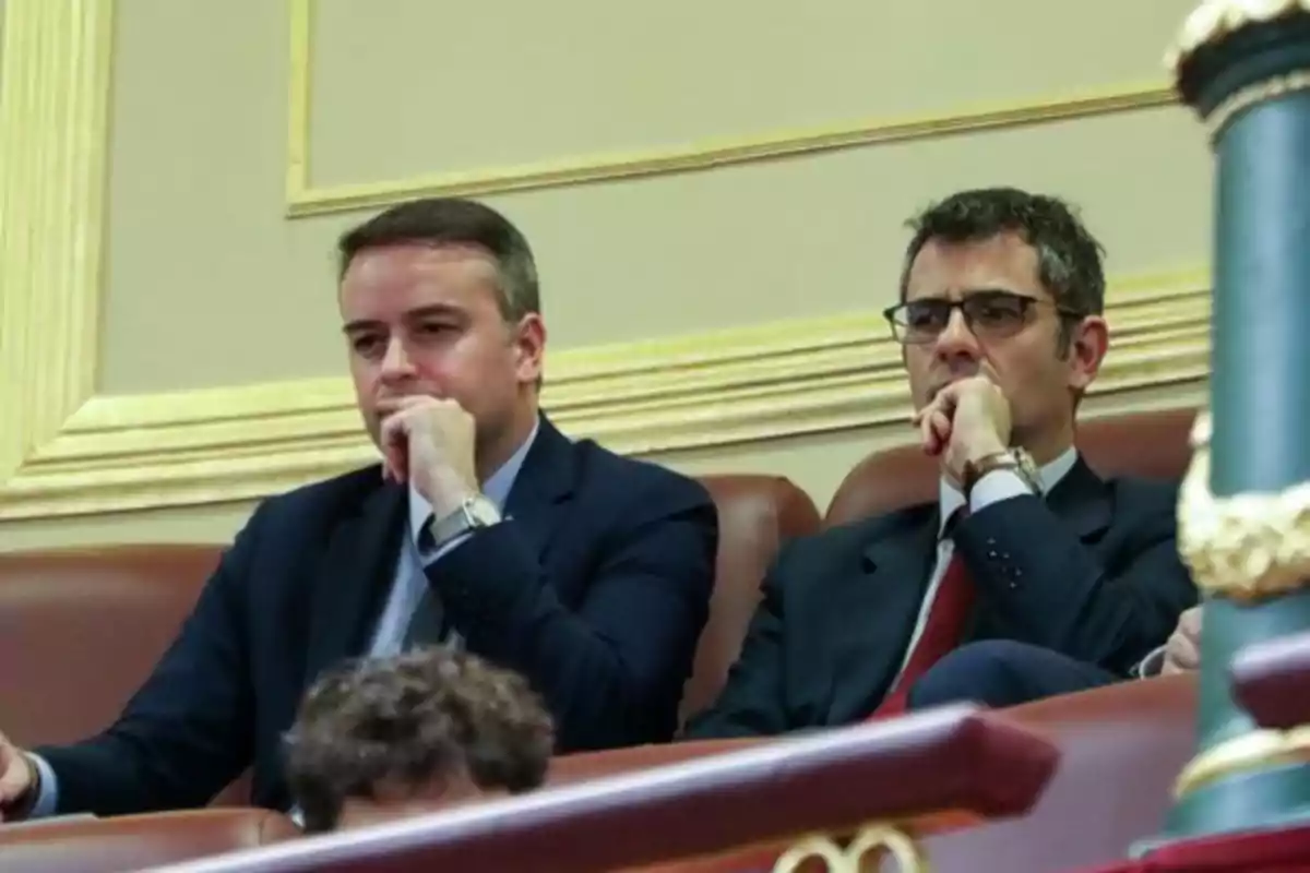 Dos hombres con traje y corbata sentados en un recinto parlamentario, ambos con expresión seria y la mano en el rostro en actitud pensativa Dos hombres con traje y corbata sentados en un recinto parlamentario, ambos con expresión seria y la mano en el rostro en actitud pensativa