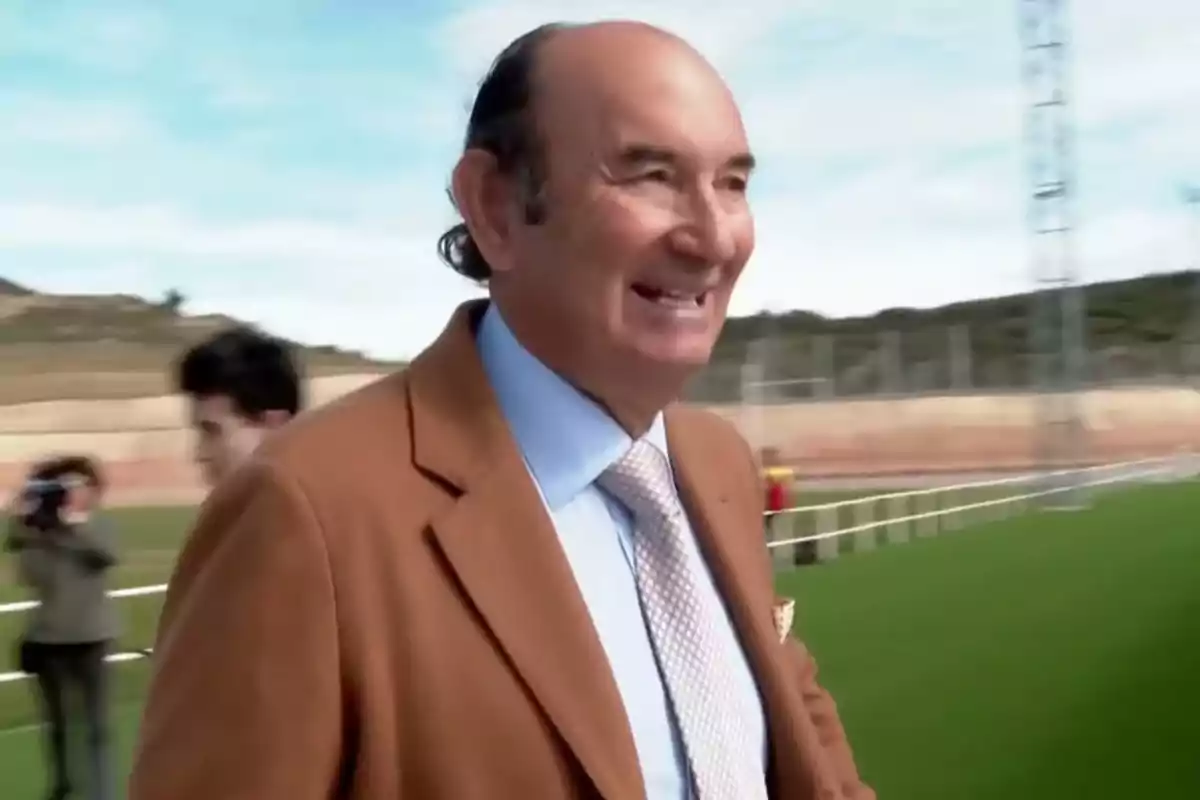 Hombre sonriente con saco marrón y corbata clara caminando en un campo deportivo al aire libre Hombre sonriente con saco marrón y corbata clara caminando en un campo deportivo al aire libre