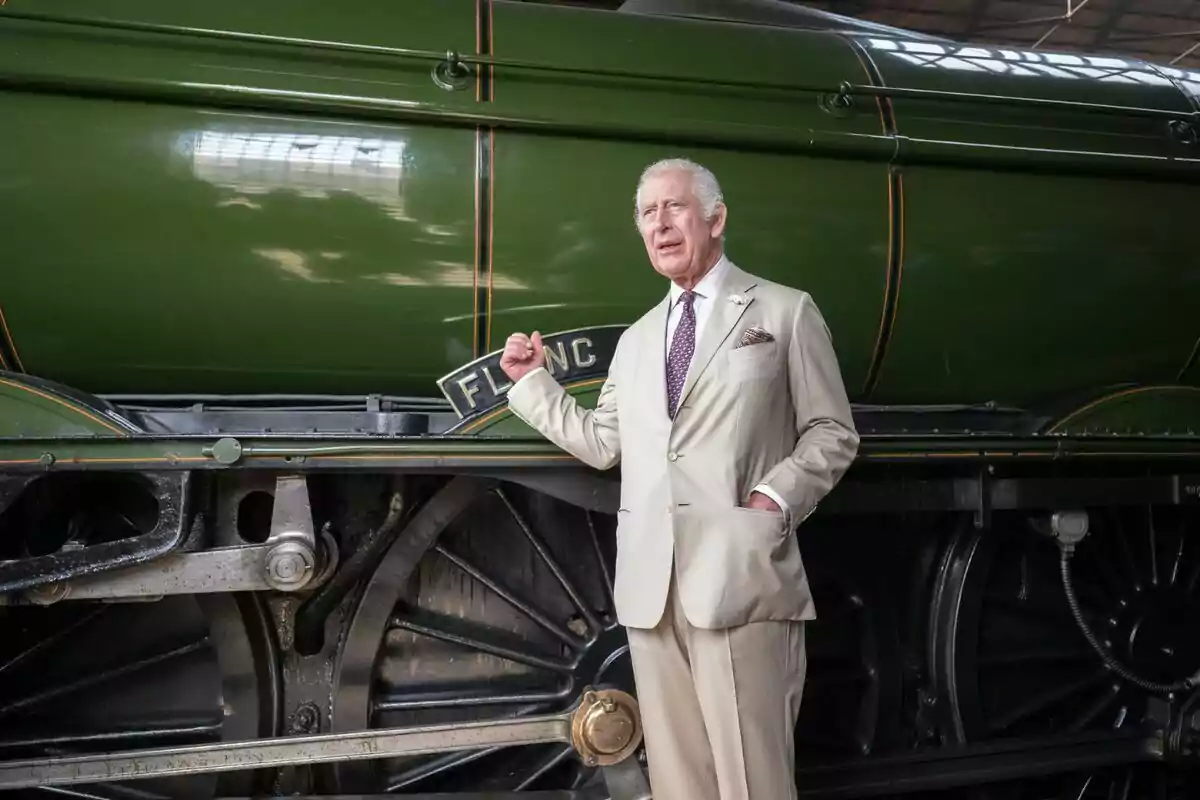 Carlos III con cabello canoso y con traje beige posando junto a una locomotora verde antigua. Carlos III con cabello canoso y con traje beige posando junto a una locomotora verde antigua.