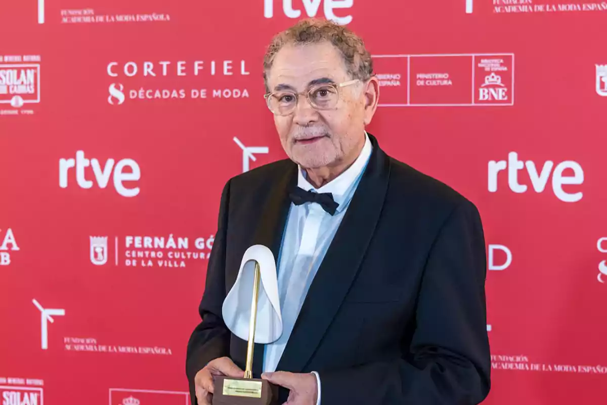 Hombre mayor con gafas y esmoquin sosteniendo un trofeo frente a un fondo rojo con logotipos de patrocinadores. Hombre mayor con gafas y esmoquin sosteniendo un trofeo frente a un fondo rojo con logotipos de patrocinadores.
