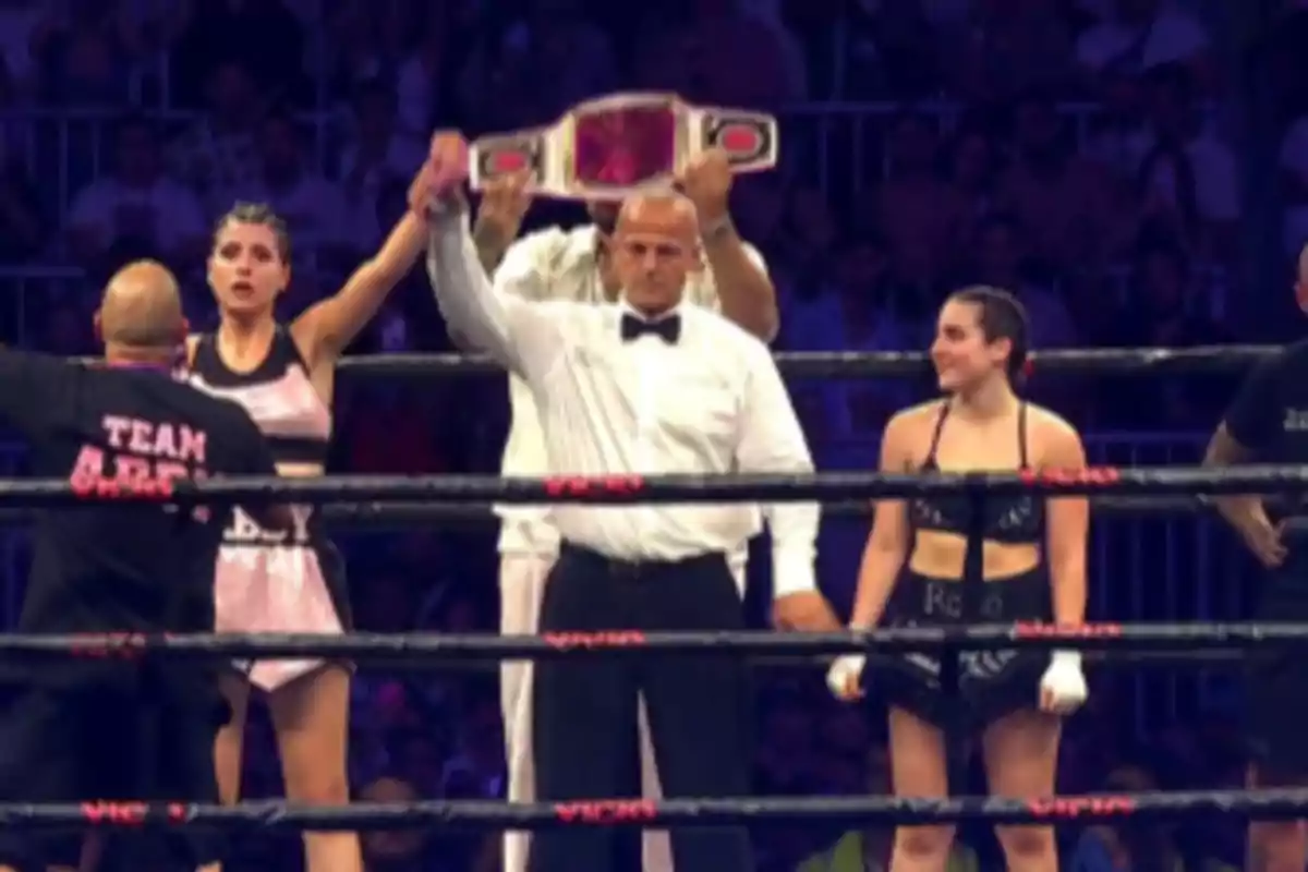 Dos boxeadoras en el ring junto a un árbitro que levanta el brazo de la ganadora mientras una persona sostiene un cinturón de campeonato al fondo Dos boxeadoras en el ring junto a un árbitro que levanta el brazo de la ganadora mientras una persona sostiene un cinturón de campeonato al fondo