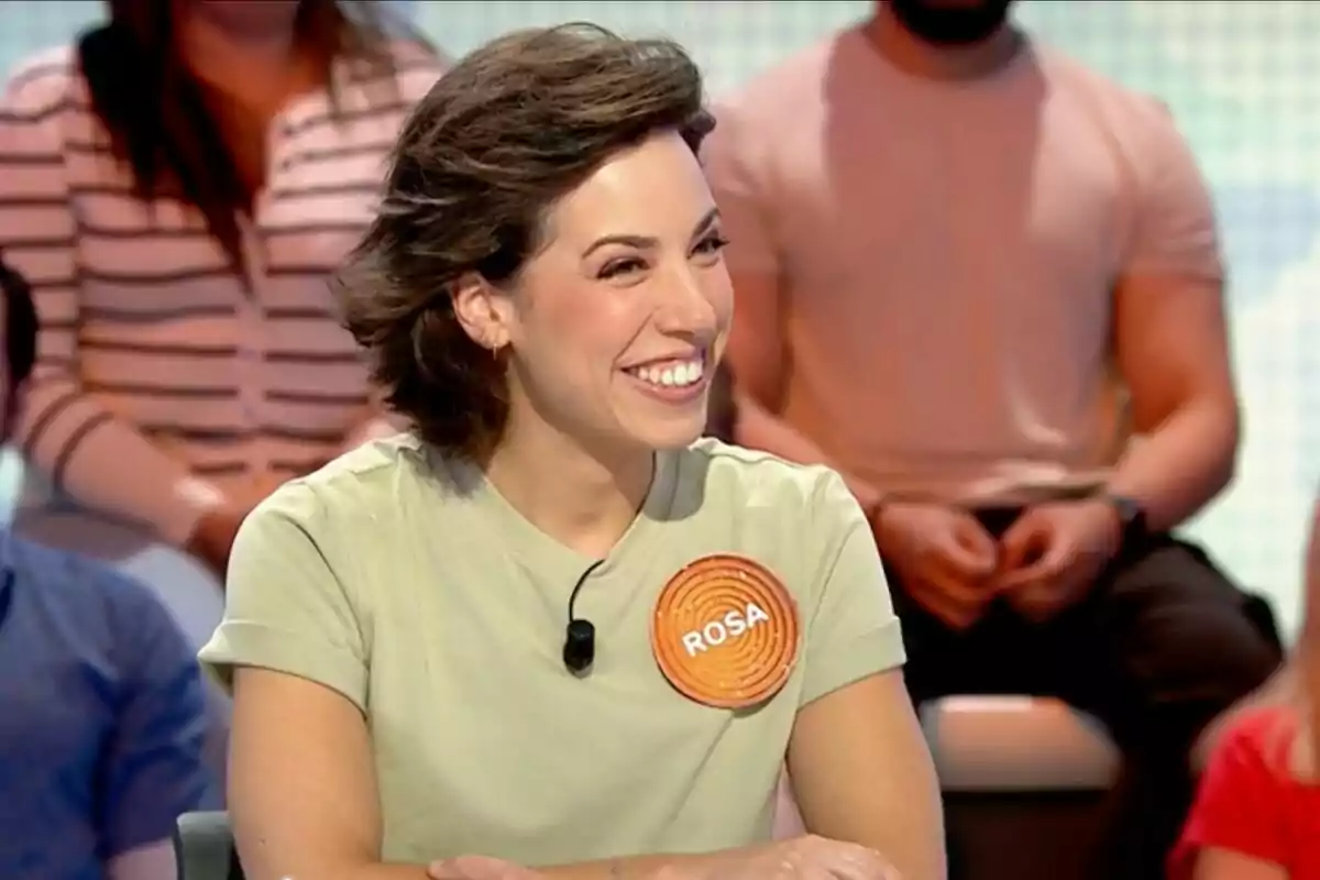 Rosa Rodríguez sonriente sentada en un plató de televisión, que es el de 'Pasapalabra', con una camiseta clara y un micrófono, lleva una chapa con el nombre Rosa y hay personas desenfocadas al fondo. Rosa Rodríguez sonriente sentada en un plató de televisión, que es el de 'Pasapalabra', con una camiseta clara y un micrófono, lleva una chapa con el nombre Rosa y hay personas desenfocadas al fondo.