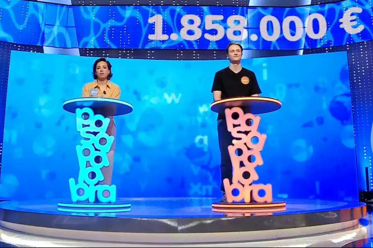 Dos concursantes, que son Rosa y Manu, de pie en sus respectivos atriles en el set del programa de televisión 'Pasapalabra' con un marcador grande que muestra un premio de un millón ochocientos cincuenta y ocho mil euros en el fondo azul. Dos concursantes, que son Rosa y Manu, de pie en sus respectivos atriles en el set del programa de televisión 'Pasapalabra' con un marcador grande que muestra un premio de un millón ochocientos cincuenta y ocho mil euros en el fondo azul.